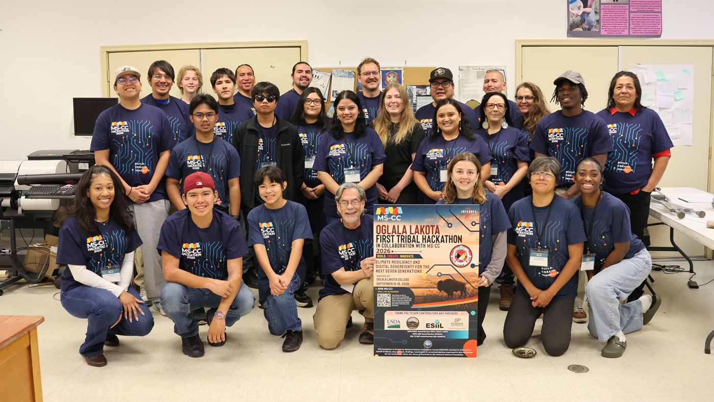 A large group of participants poses together wearing matching blue MS-CC t-shirts, with a man kneeling in front holding an event poster for the Oglala Lakota First Tribal Hackathon.