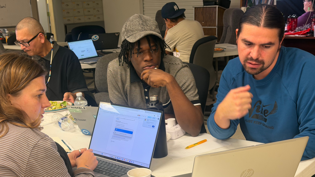 Three team members sit together at a table focused on a laptop screen, discussing their work during a collaborative session.
